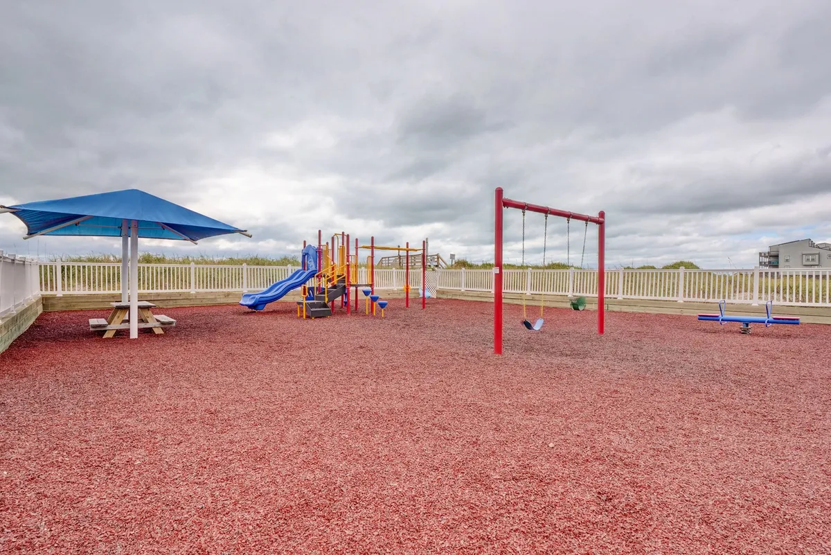 A lively outdoor playground with colorful equipment, swings, and a nearby picnic area, all surrounded by safety fencing and greenery. Children are playing while families gather for picnics and social interaction.