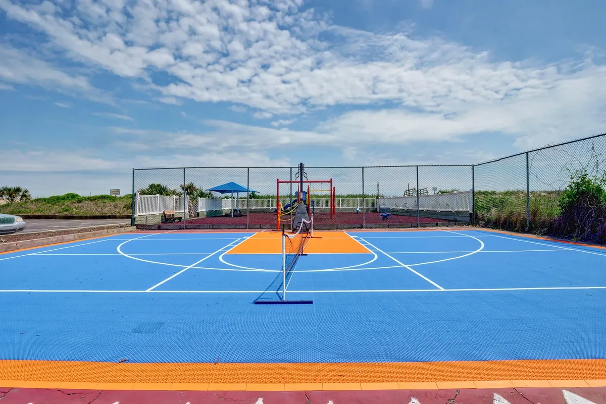 A colorful basketball court set outdoors, complete with netting for basketball or tennis, is surrounded by lush greenery. The vibrant court surface and open sky create a playful, energetic atmosphere, encouraging friendly family competitions and active play.