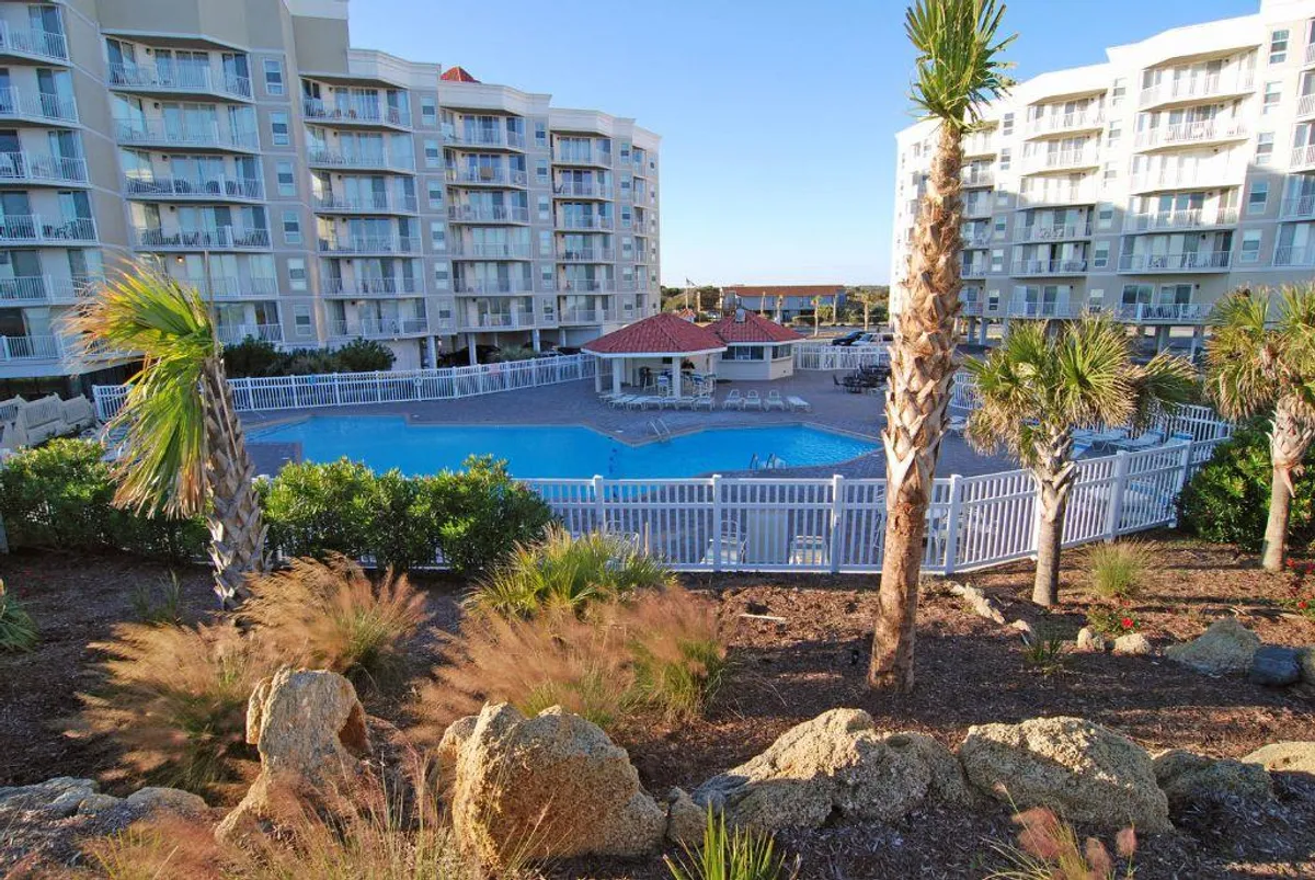 This exterior image showcases a sparkling swimming pool surrounded by palm trees, comfortable lounge chairs, and the resort's modern apartment building with a distinctive clubhouse featuring a red roof. The tranquil ambiance is enhanced by the leisurely setting and hints of sunset lighting, evoking relaxation and wellness.