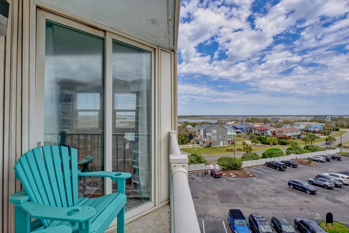 A tranquil private balcony with a turquoise chair positioned to face sweeping ocean views. The vibrant blue sea stretches to the horizon, while colorful houses dot the shoreline below. The atmosphere suggests early morning light or the golden hues of sunset, promising peaceful moments at any time of day.