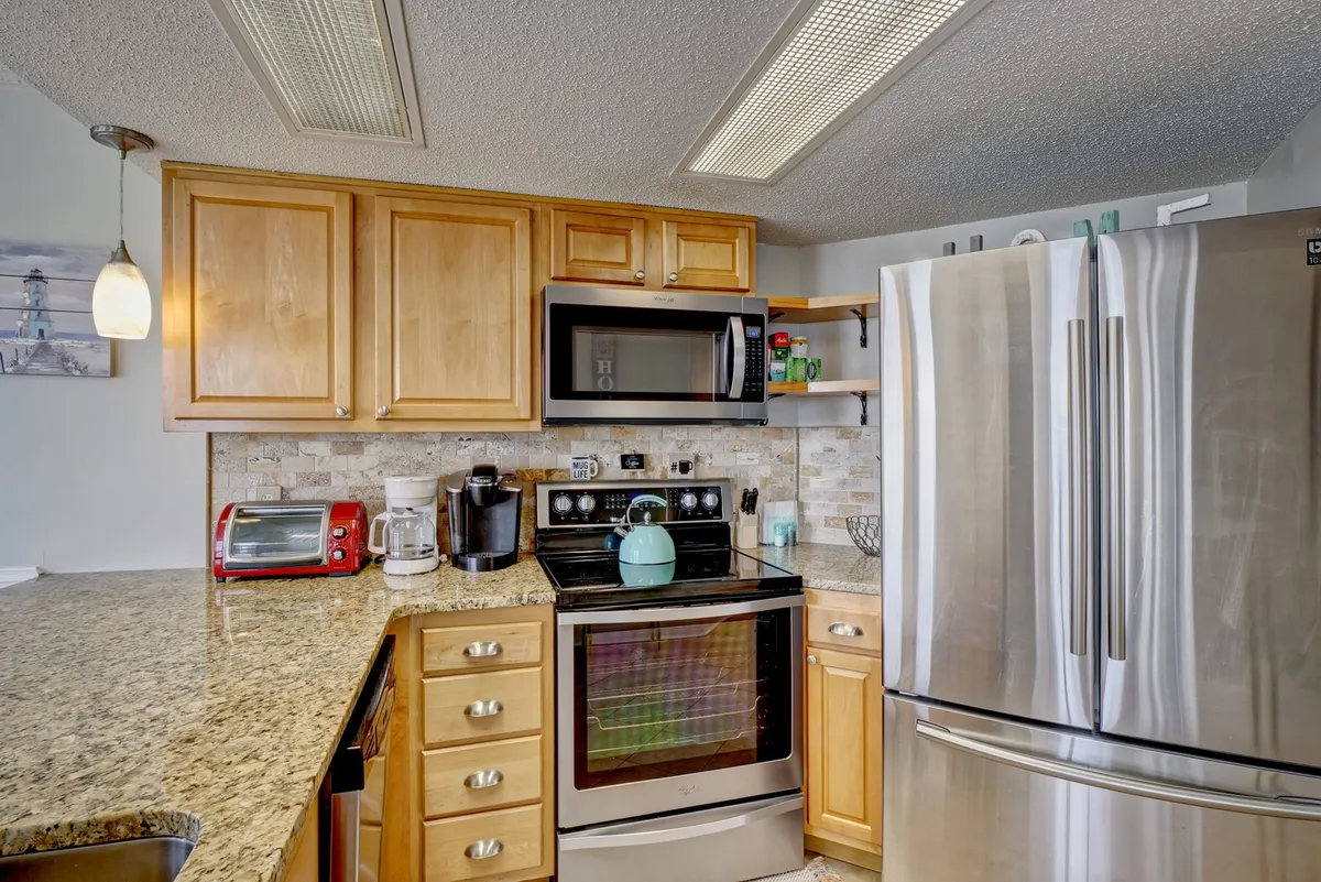 This image spotlights the kitchen area, clearly displaying granite countertops and stainless steel appliances. A teal kettle and decorative signage add a welcoming touch, while the cabinetry and ambient lighting reinforce the modern, beach-chic vibe described in the section.