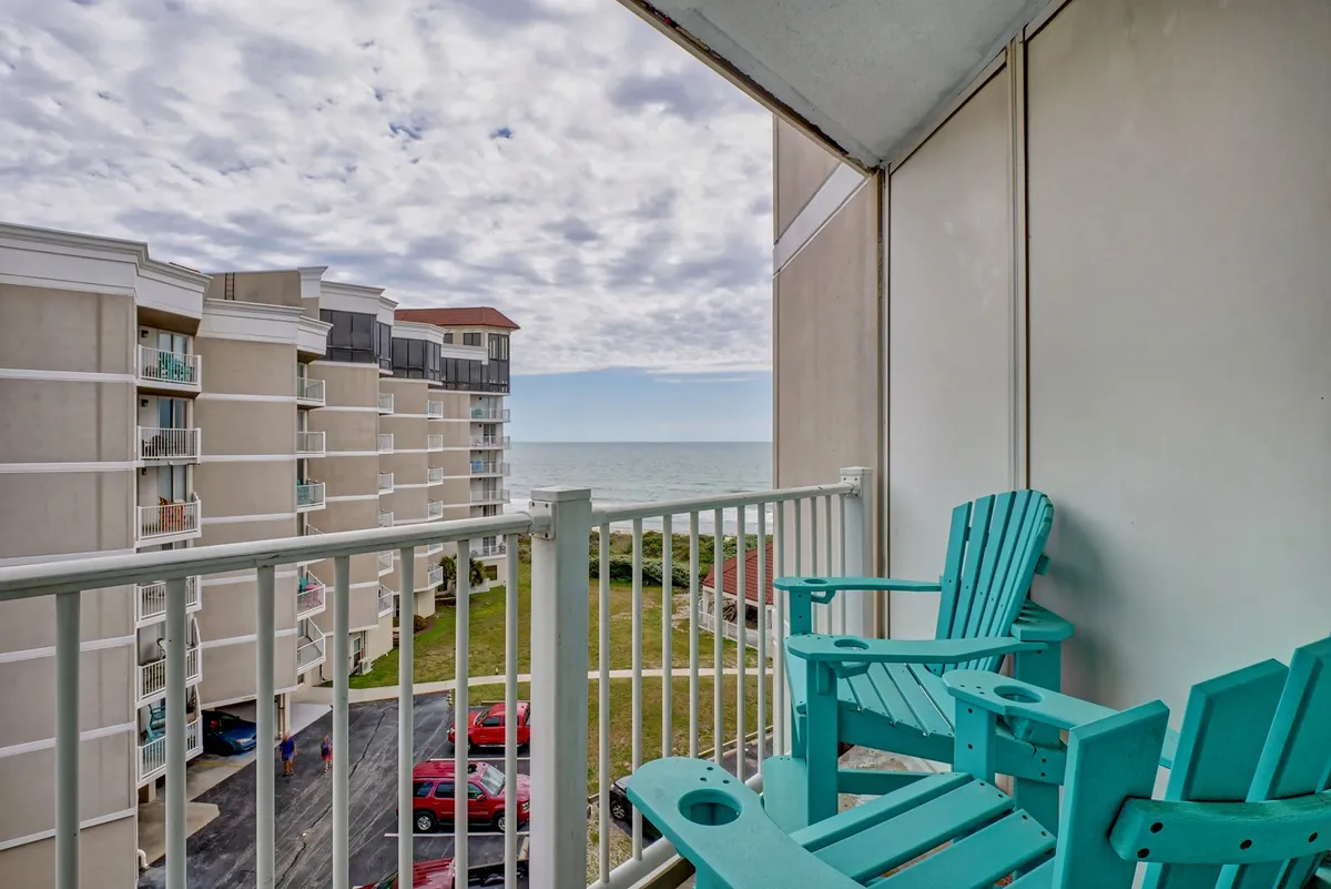 The image showcases an inviting balcony with Adirondack chairs overlooking an expansive ocean view. The scene captures the tranquility of the coastline with the sun rising over the water, highlighting the perfect spot for relaxation and quiet enjoyment.