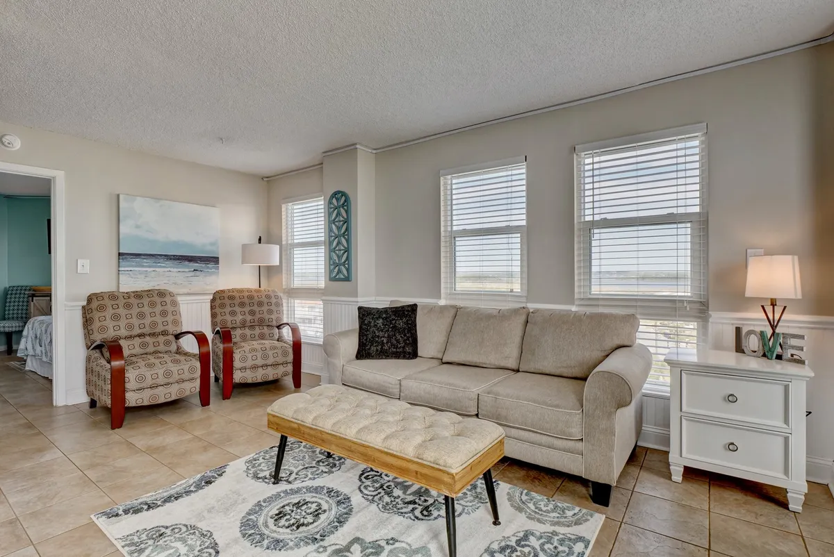 A comfortable living space just inside, featuring a sofa, armchairs, a coffee table, and tasteful coastal artwork. Large windows suggest a connection to the balcony and the outdoors, with soft daylight filling the room and inviting guests to step outside.