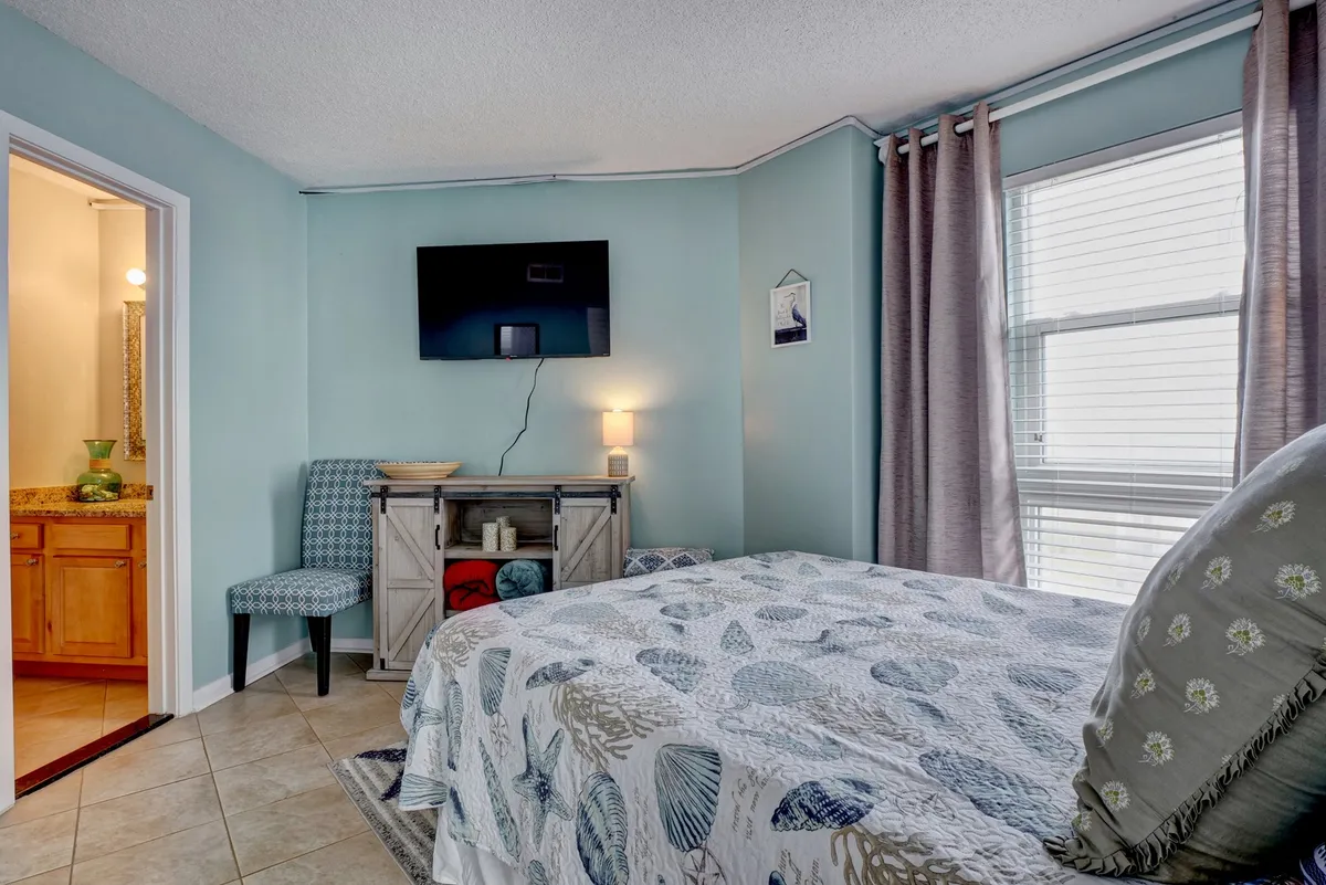 A serene primary bedroom featuring a large bed with patterned bedding, a wall-mounted television, a decorative chair, and a view into the connected bathroom. Natural sunlight streams in, creating a restful and inviting space.