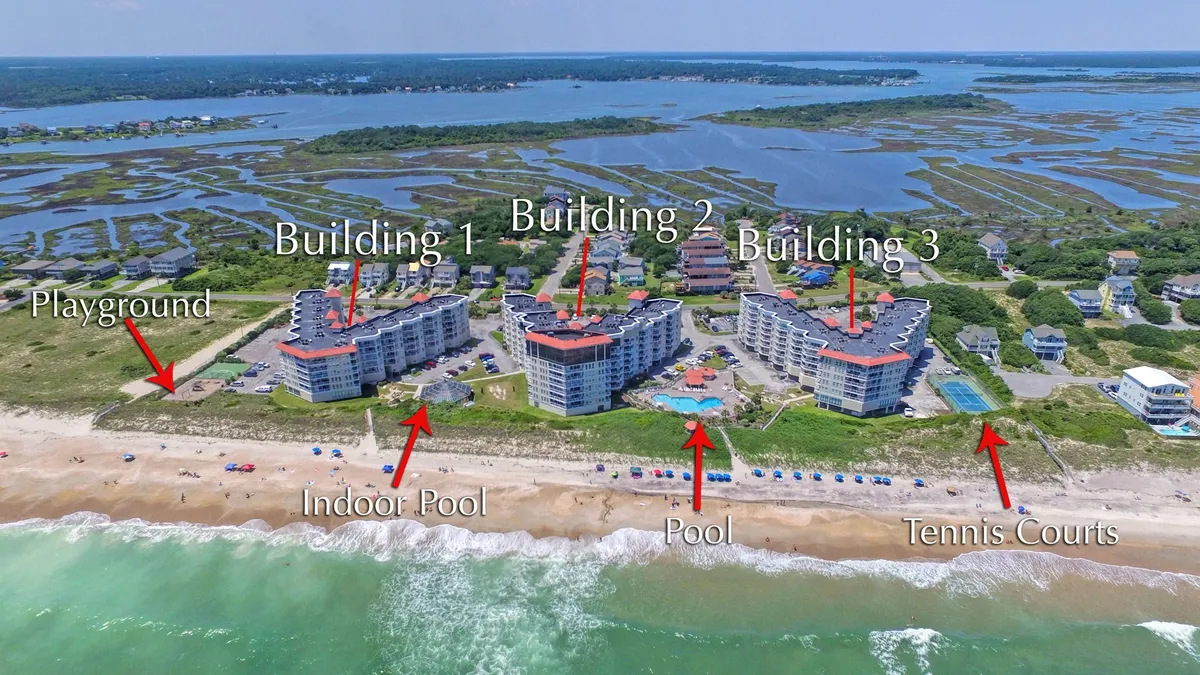 A wide aerial view showing both the indoor and outdoor swimming pools, vibrant tennis courts, a children's playground, and nearby beach area, all surrounded by red-roofed buildings and lush greenery. Families and guests can be seen enjoying various amenities, from swimming and sports to relaxing by the water.