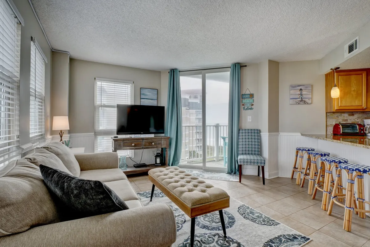 This image highlights the comfortable living area with a sofa, television, and large sliding balcony doors that flood the space with natural light. Kitchen bar stools are visible in the background, reinforcing the connection between living and dining zones.