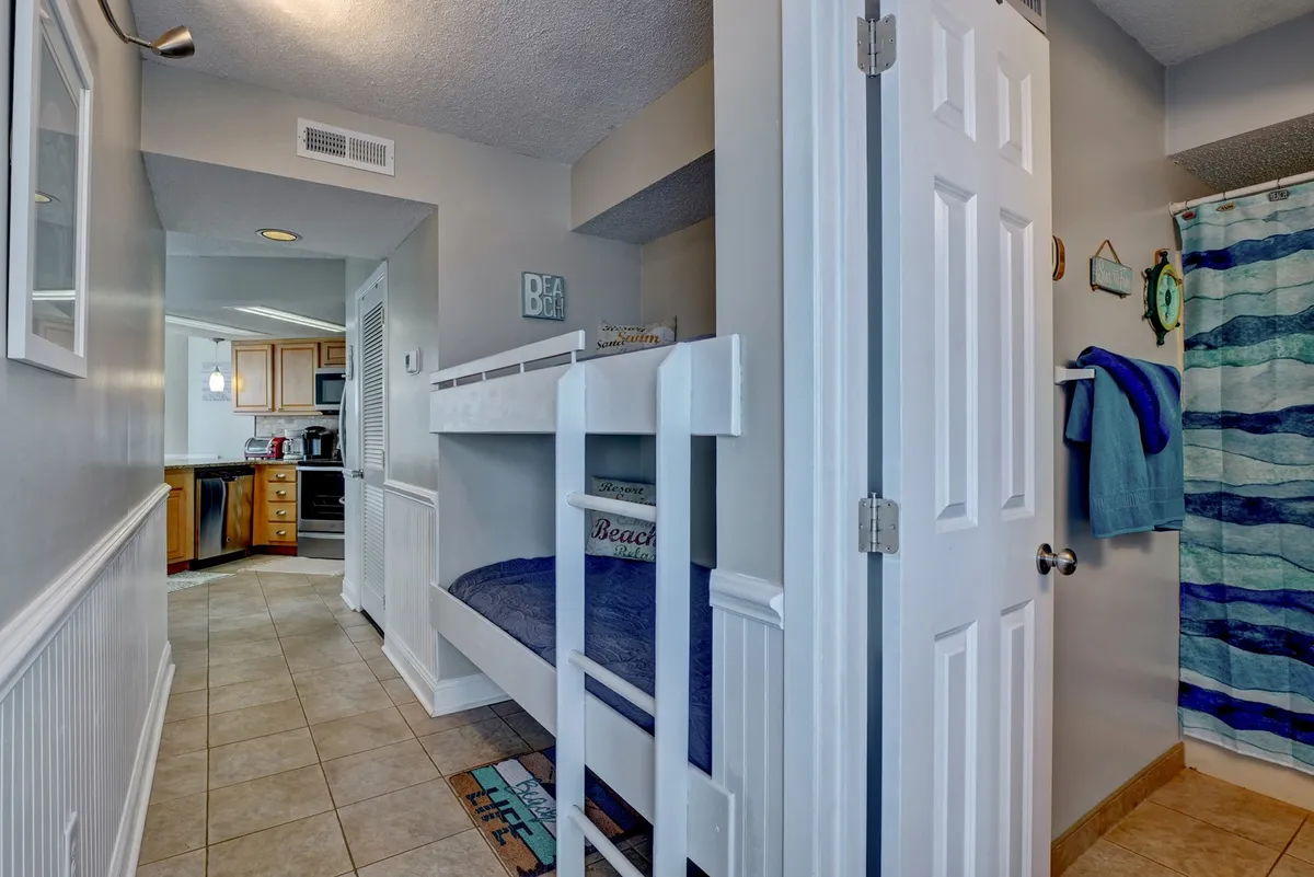 A playful hallway nook with built-in bunk beds featuring beach-themed decor, adjacent to the kitchen entrance and bathroom door. The design maximizes space and creates a cozy sleeping spot for young guests.